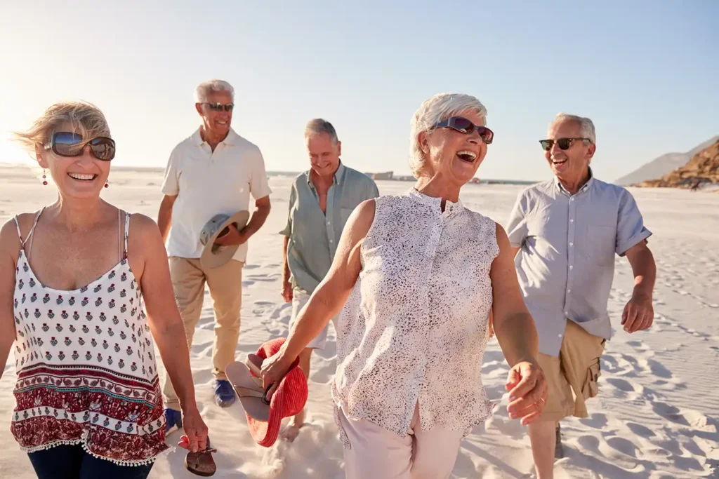 Portrait Of Senior Friends Standing On Rocks By Sea On Summer Group Vacation With Arms Outstretched